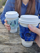 Load image into Gallery viewer, teal drink cozie with feather print drink cozie on a cup being held by someone beside a blue drink cozie on a cup being held by another
