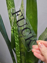 Load image into Gallery viewer, Clear 2 by 6 inch arcylic bookmark with silky tassel on a metal chain black FUCK. design in front of a snake plant 
